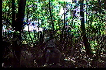 Back in woods, seated on same rock, holding up new rifle